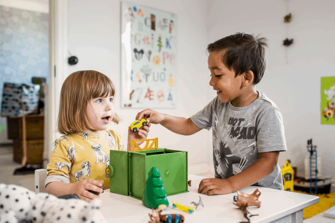 Two young children play with toy cars and animals at a table in a brightly decorated room, smiling and interacting with each other.